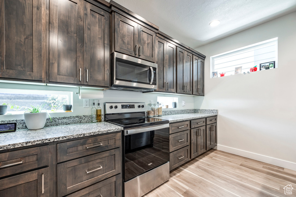 Kitchen with appliances with stainless steel finishes, dark brown cabinetry, baseboards, light wood-style floors, and light stone countertops