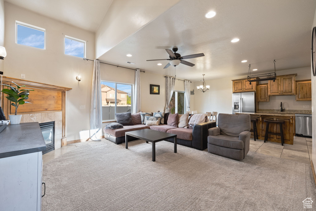 Carpeted living room with a high ceiling, a tiled fireplace, ceiling fan with notable chandelier, and sink