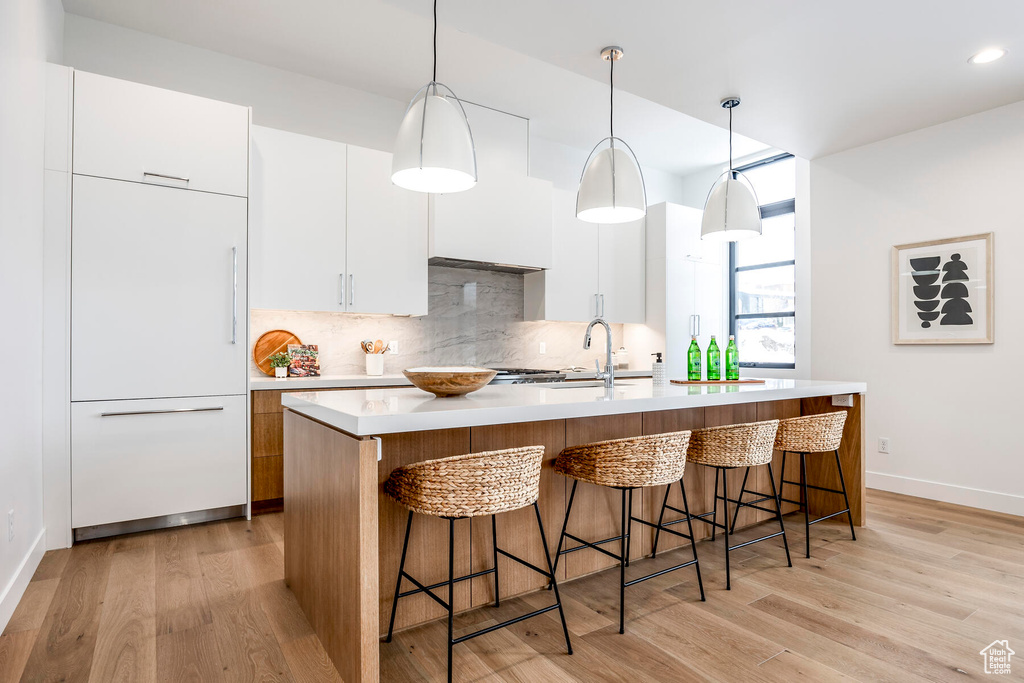 Kitchen with light wood-type flooring, white cabinetry, backsplash, an island with sink, and hanging light fixtures