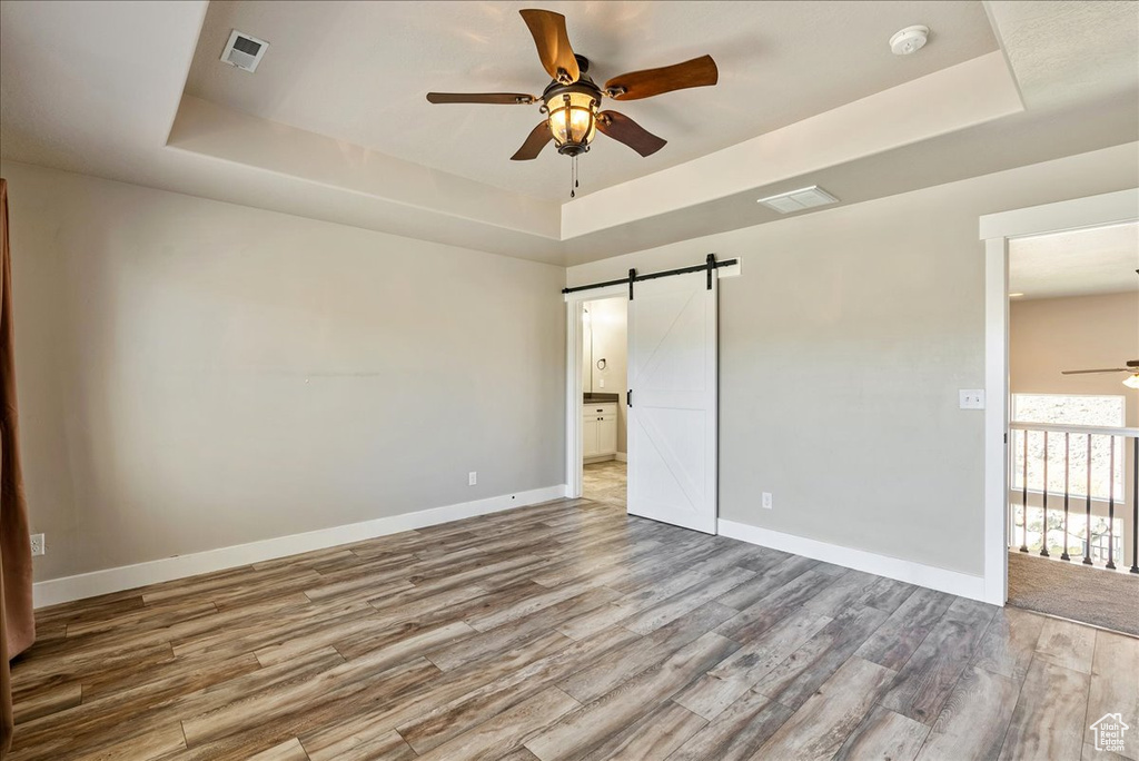 Unfurnished bedroom with ceiling fan, a raised ceiling, a barn door, and wood-type flooring