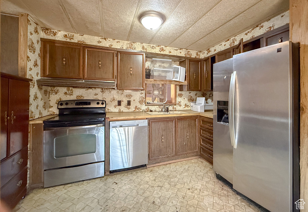 Kitchen featuring stainless steel appliances, light countertops, brown cabinetry, a sink, and wallpapered walls