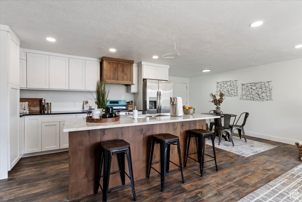 Kitchen featuring an island with sink, stainless steel fridge, dark wood-type flooring, and white cabinetry