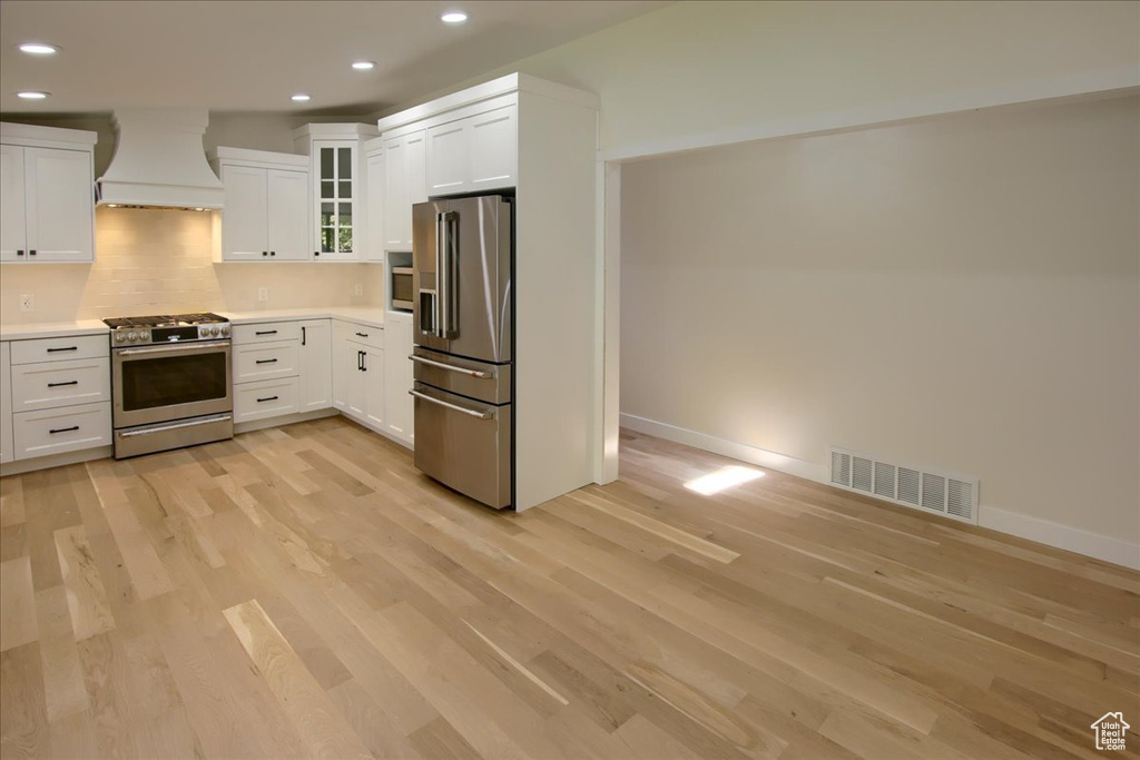 Kitchen featuring premium range hood, tasteful backsplash, light hardwood / wood-style flooring, white cabinetry, and appliances with stainless steel finishes