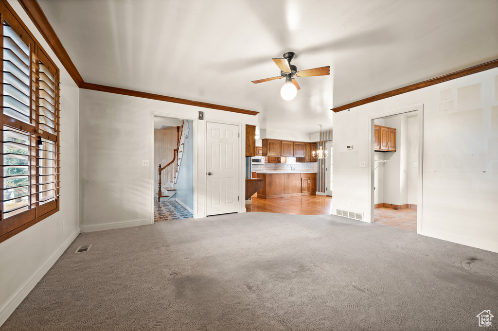 Unfurnished living room featuring light carpet, ceiling fan with notable chandelier, and ornamental molding