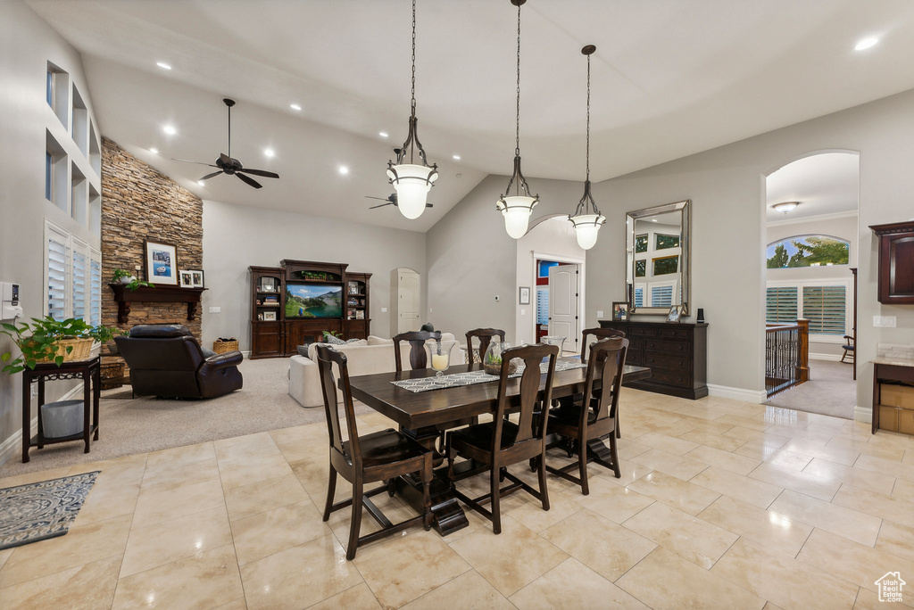 Dining area with ceiling fan, a stone fireplace, light colored carpet, and high vaulted ceiling