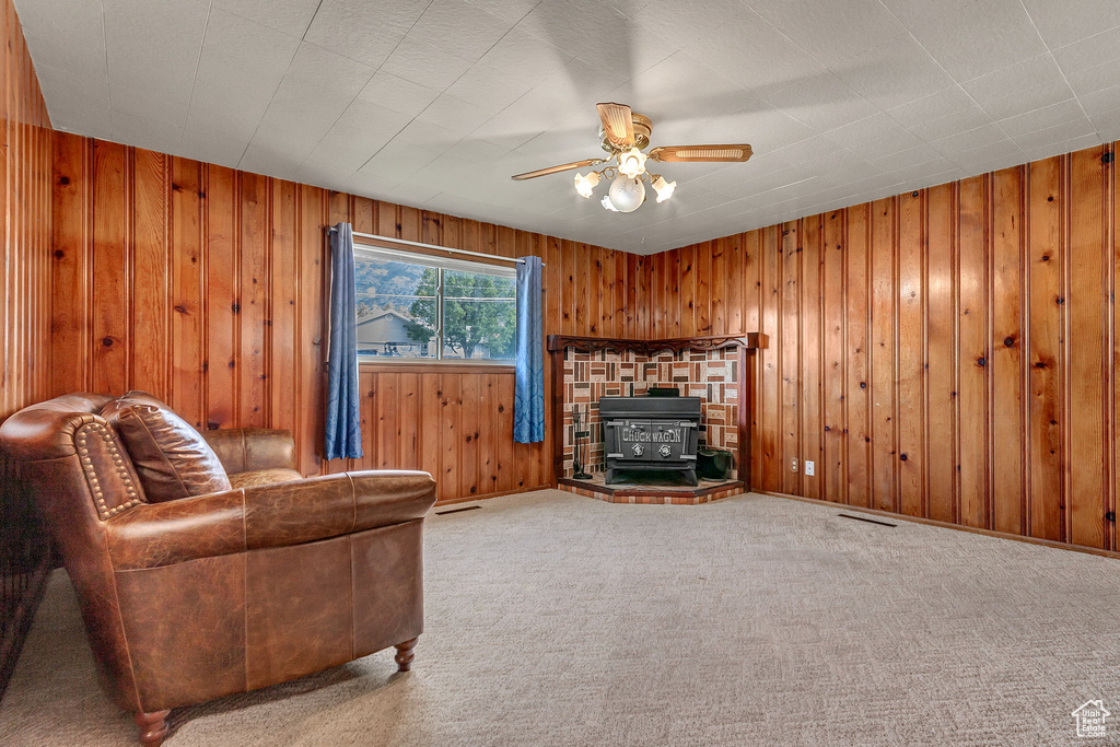 Carpeted living room featuring wooden walls, ceiling fan, and a wood stove