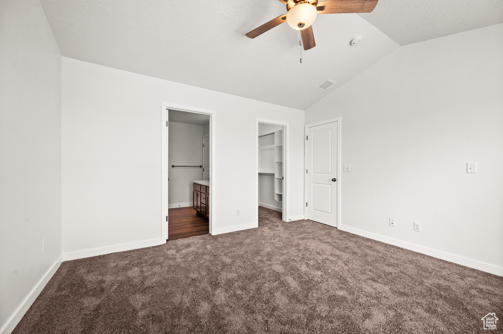 Unfurnished bedroom featuring dark carpet, vaulted ceiling, ensuite bath, a walk in closet, and ceiling fan