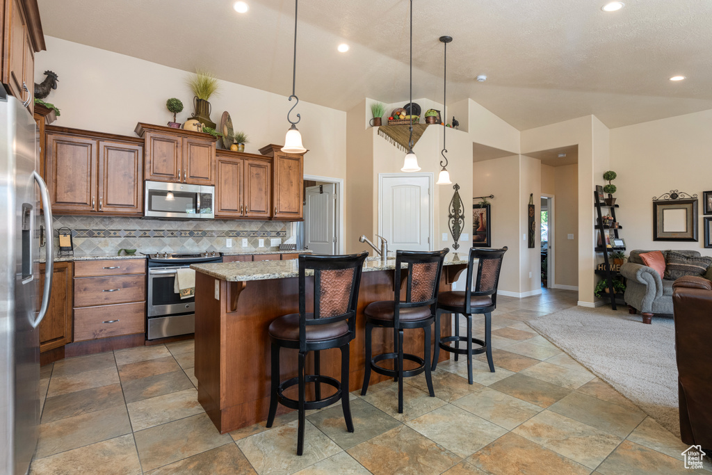 Kitchen featuring an island with sink, decorative light fixtures, stainless steel appliances, light stone countertops, and decorative backsplash