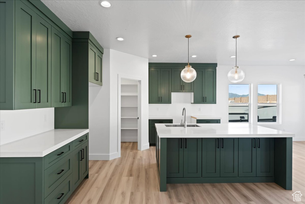 Kitchen with light wood-type flooring, sink, green cabinetry, hanging light fixtures, and a center island with sink