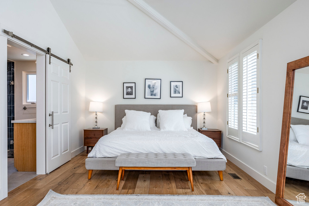 Bedroom featuring a barn door, light wood-type flooring, lofted ceiling with beams, and ensuite bath