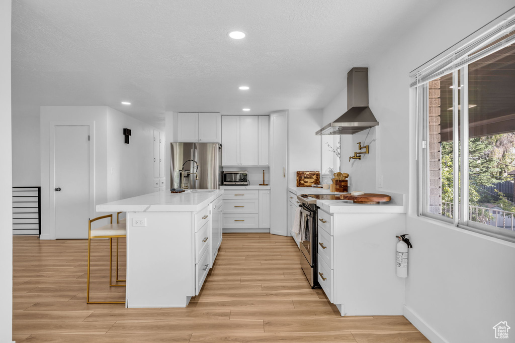 Kitchen featuring white cabinetry, a kitchen bar, wall chimney exhaust hood, stainless steel appliances, and light hardwood / wood-style flooring