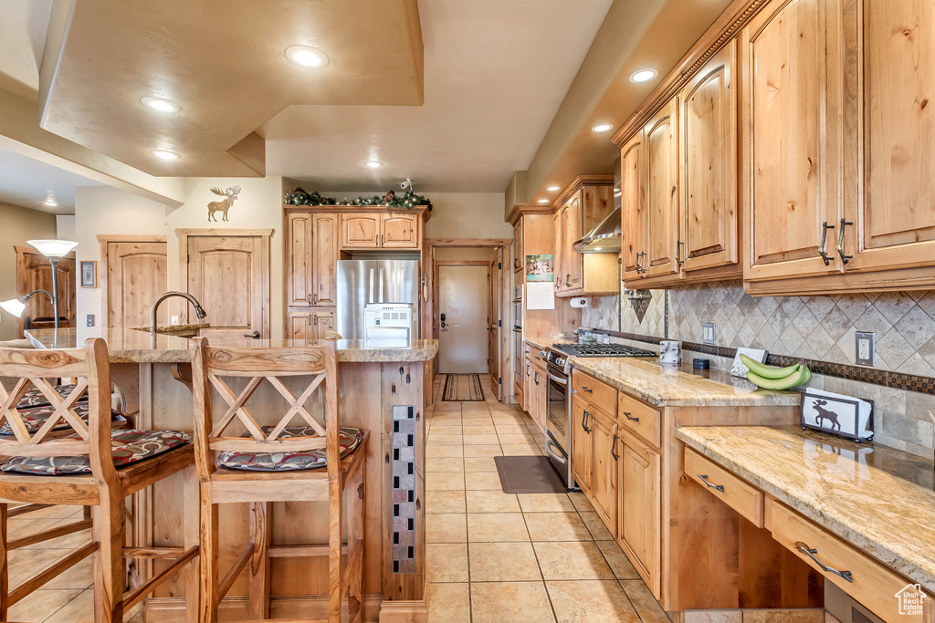 Kitchen featuring light stone counters, tasteful backsplash, a kitchen breakfast bar, appliances with stainless steel finishes, and light tile patterned floors