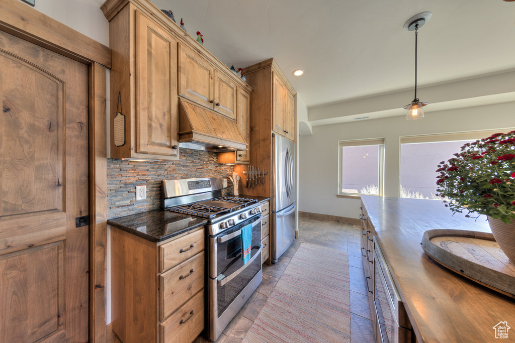 Kitchen with appliances with stainless steel finishes, decorative backsplash, hanging light fixtures, dark stone counters, and premium range hood