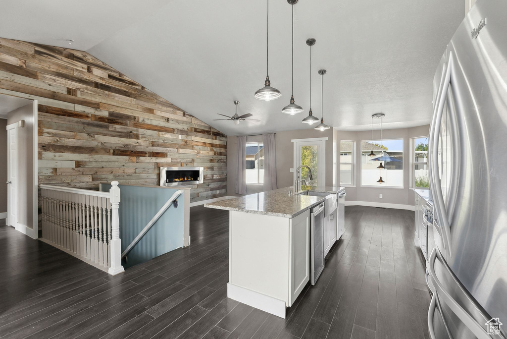 Kitchen with an island with sink, hanging light fixtures, wood walls, stainless steel refrigerator, and light stone countertops