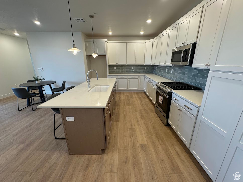 Kitchen with a kitchen island with sink, stainless steel appliances, white cabinets, sink, and hanging light fixtures