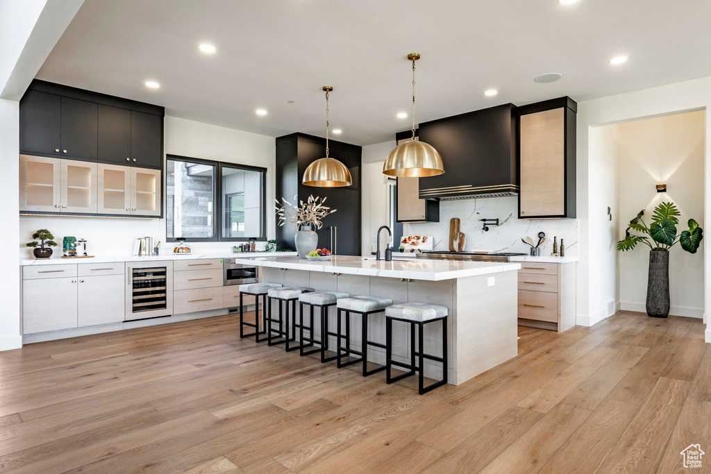 Kitchen featuring light wood-type flooring, an island with sink, beverage cooler, hanging light fixtures, and exhaust hood