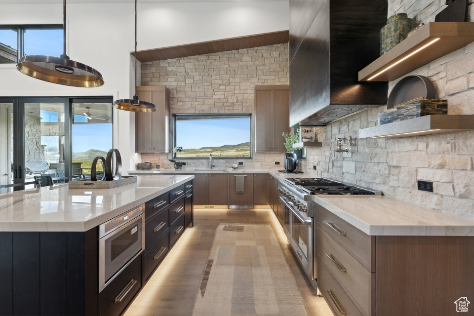 Kitchen with decorative light fixtures, a kitchen island with sink, backsplash, appliances with stainless steel finishes, and light stone countertops