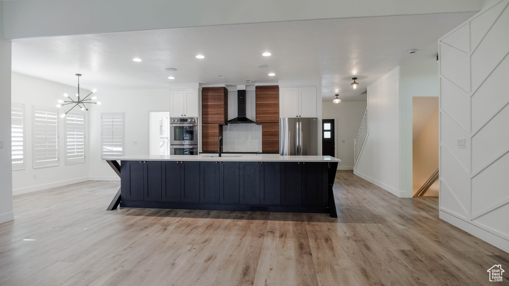 Kitchen featuring wall chimney exhaust hood, a large island with sink, stainless steel appliances, and white cabinets