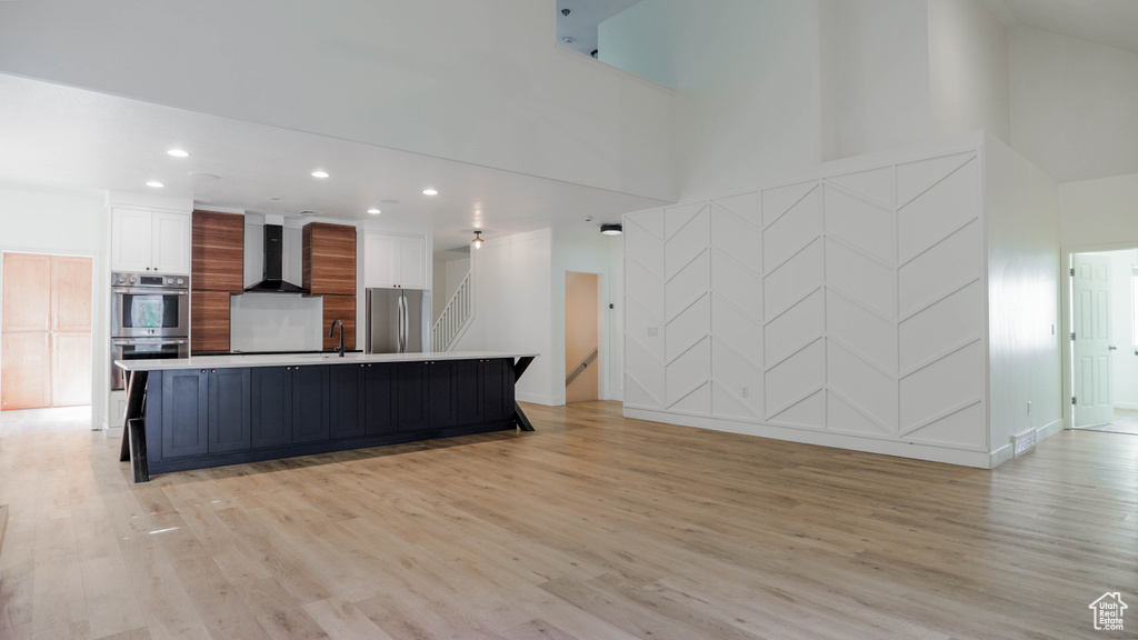 Kitchen with appliances with stainless steel finishes, a towering ceiling, white cabinets, a large island, and wall chimney range hood