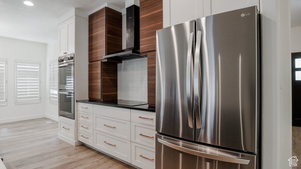 Kitchen with tasteful backsplash, light hardwood / wood-style flooring, wall chimney range hood, white cabinetry, and appliances with stainless steel finishes