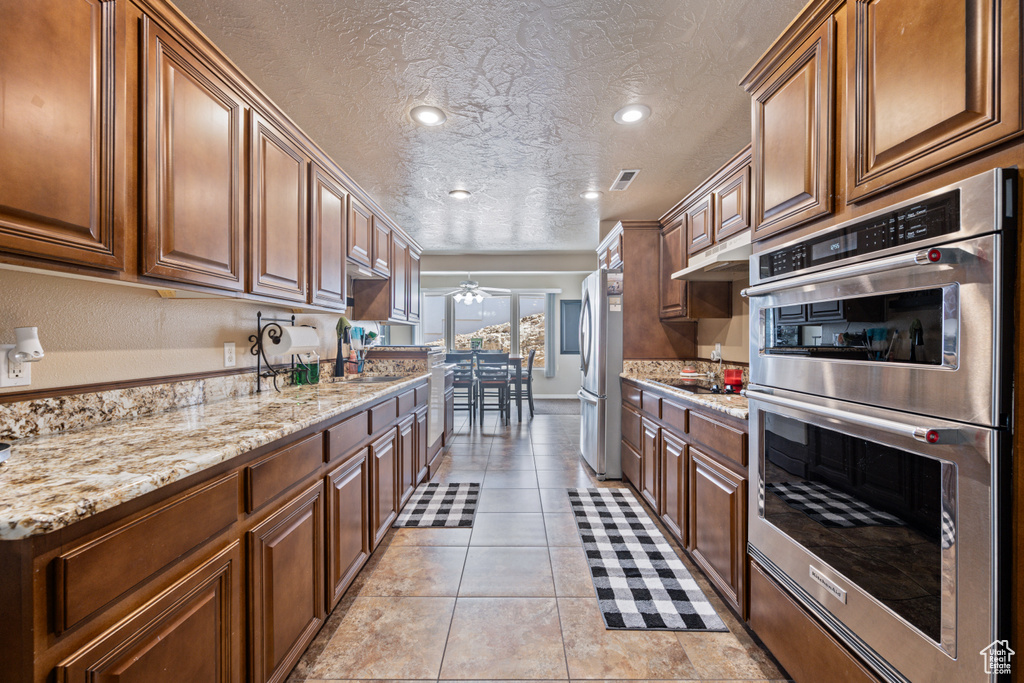 Kitchen with a textured ceiling, appliances with stainless steel finishes, light tile patterned floors, light stone countertops, and ceiling fan