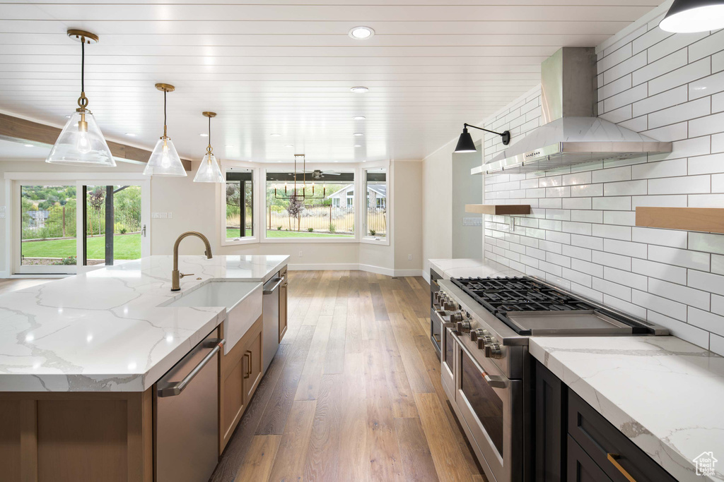 Kitchen with light stone countertops, appliances with stainless steel finishes, decorative light fixtures, and wall chimney range hood