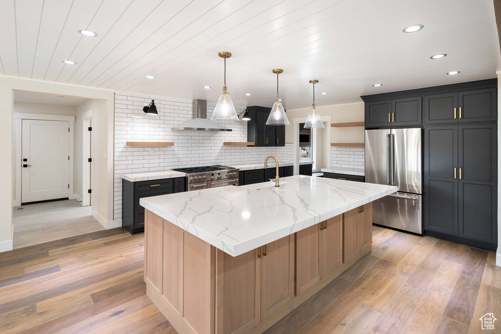 Kitchen with light stone counters, premium appliances, pendant lighting, a kitchen island with sink, and wall chimney range hood