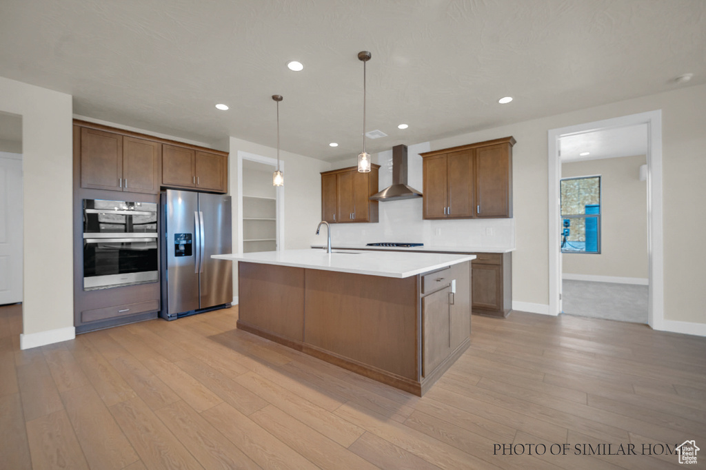 Kitchen with an island with sink, decorative light fixtures, wall chimney exhaust hood, light hardwood / wood-style flooring, and appliances with stainless steel finishes