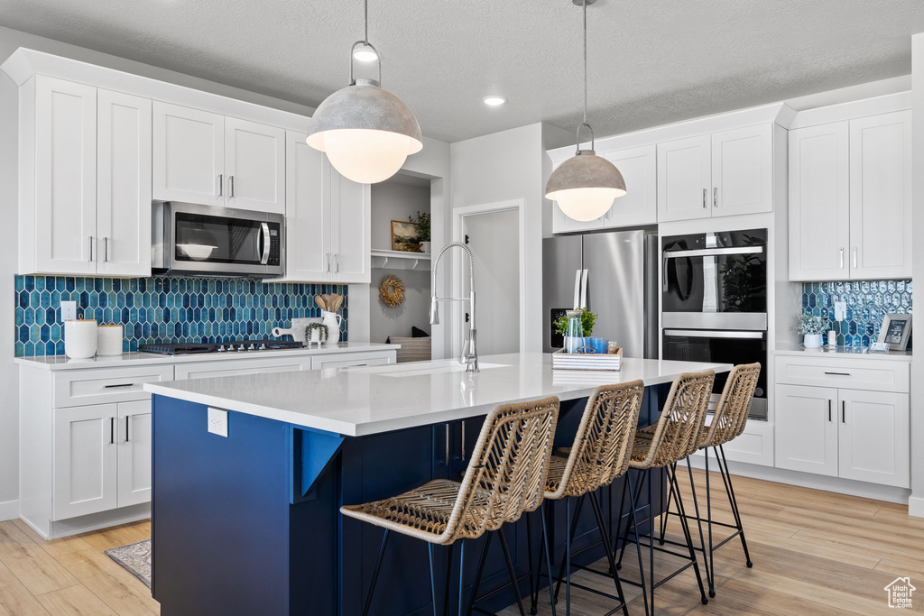 Kitchen featuring light hardwood / wood-style floors, white cabinets, hanging light fixtures, a center island with sink, and appliances with stainless steel finishes