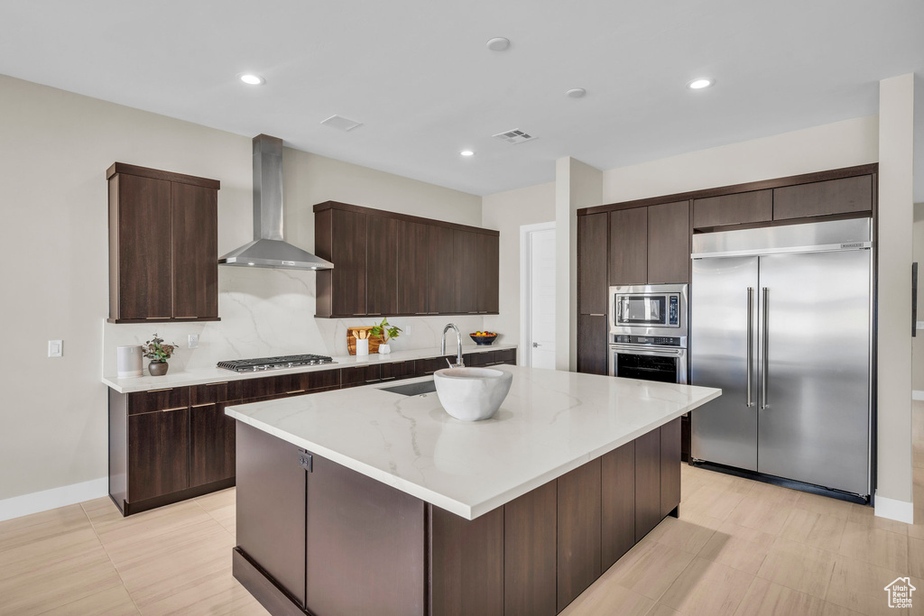 Kitchen featuring dark brown cabinetry, built in appliances, a center island with sink, and wall chimney range hood