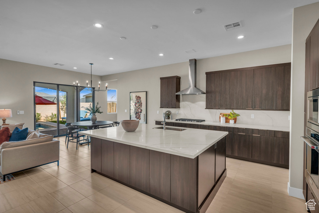 Kitchen featuring dark brown cabinetry, an island with sink, sink, wall chimney range hood, and a notable chandelier