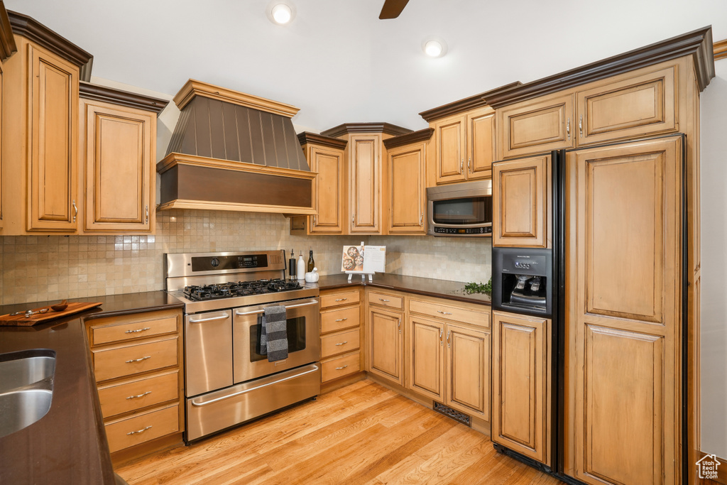 Kitchen with appliances with stainless steel finishes, backsplash, light wood-type flooring, ceiling fan, and custom exhaust hood