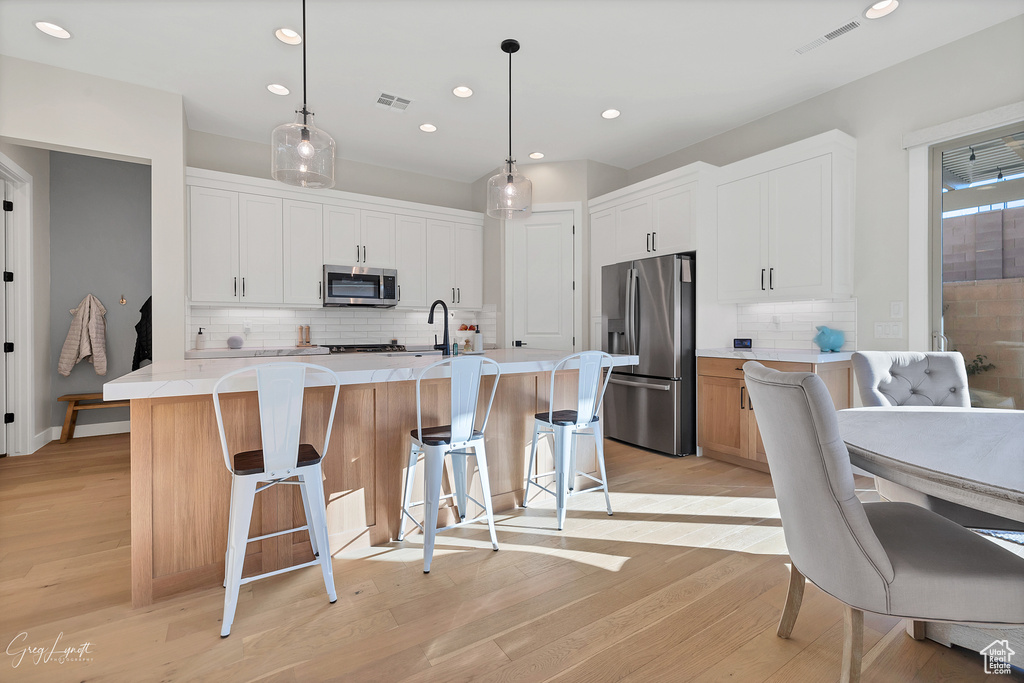 Kitchen with light hardwood / wood-style floors, white cabinets, stainless steel appliances, a center island with sink, and decorative light fixtures