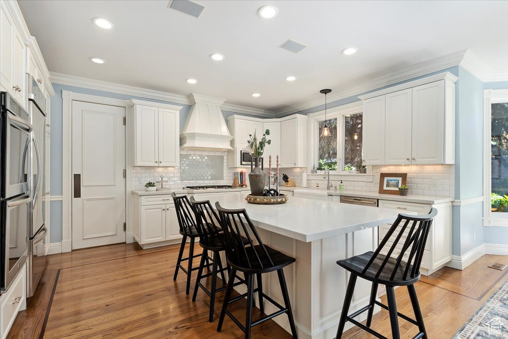 Kitchen with decorative backsplash, light hardwood / wood-style floors, white cabinetry, custom exhaust hood, and decorative light fixtures