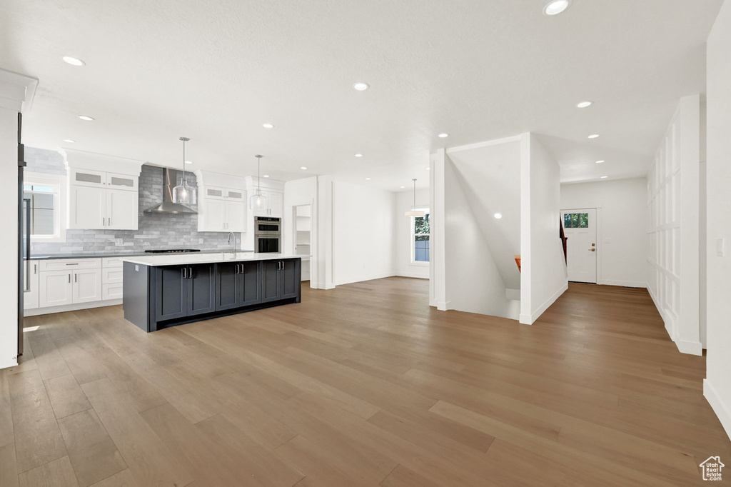 Kitchen with pendant lighting, light hardwood / wood-style floors, an island with sink, white cabinetry, and wall chimney exhaust hood