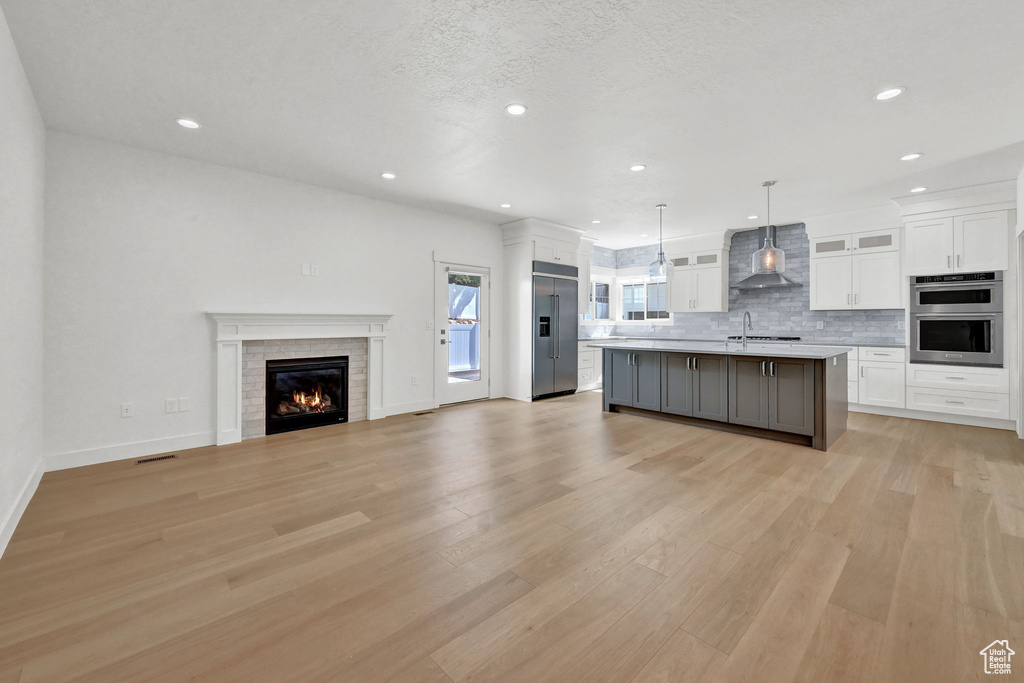 Kitchen with light hardwood / wood-style floors, a kitchen island with sink, white cabinetry, wall chimney range hood, and appliances with stainless steel finishes