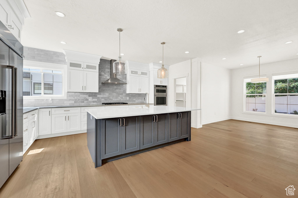 Kitchen featuring white cabinetry, light hardwood / wood-style floors, wall chimney range hood, and a center island with sink