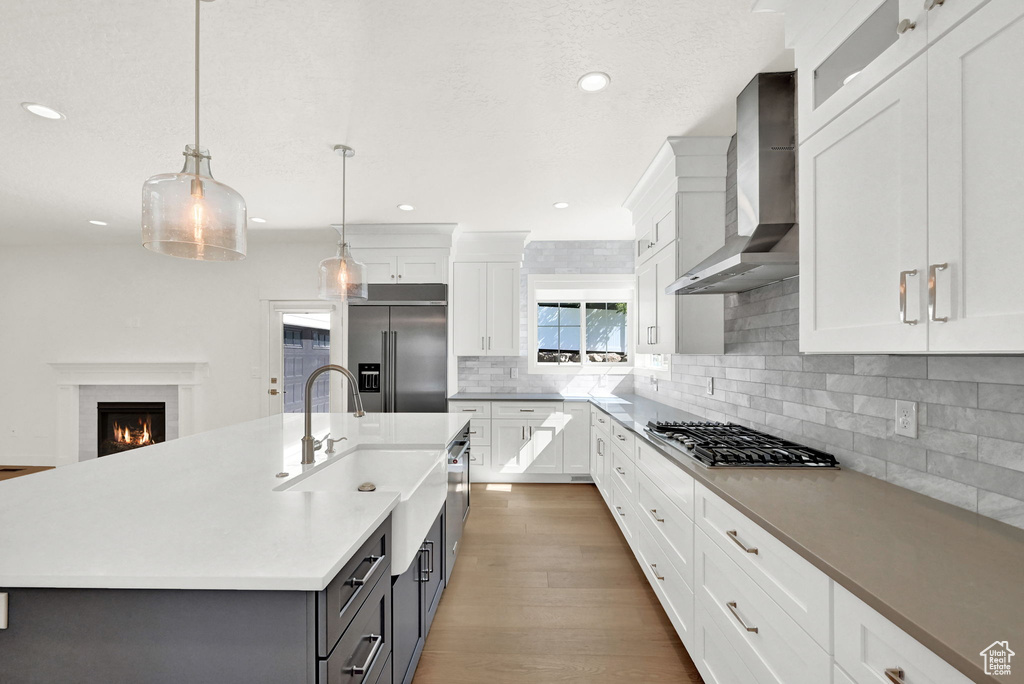 Kitchen featuring a kitchen island with sink, white cabinets, hanging light fixtures, wall chimney range hood, and stainless steel appliances