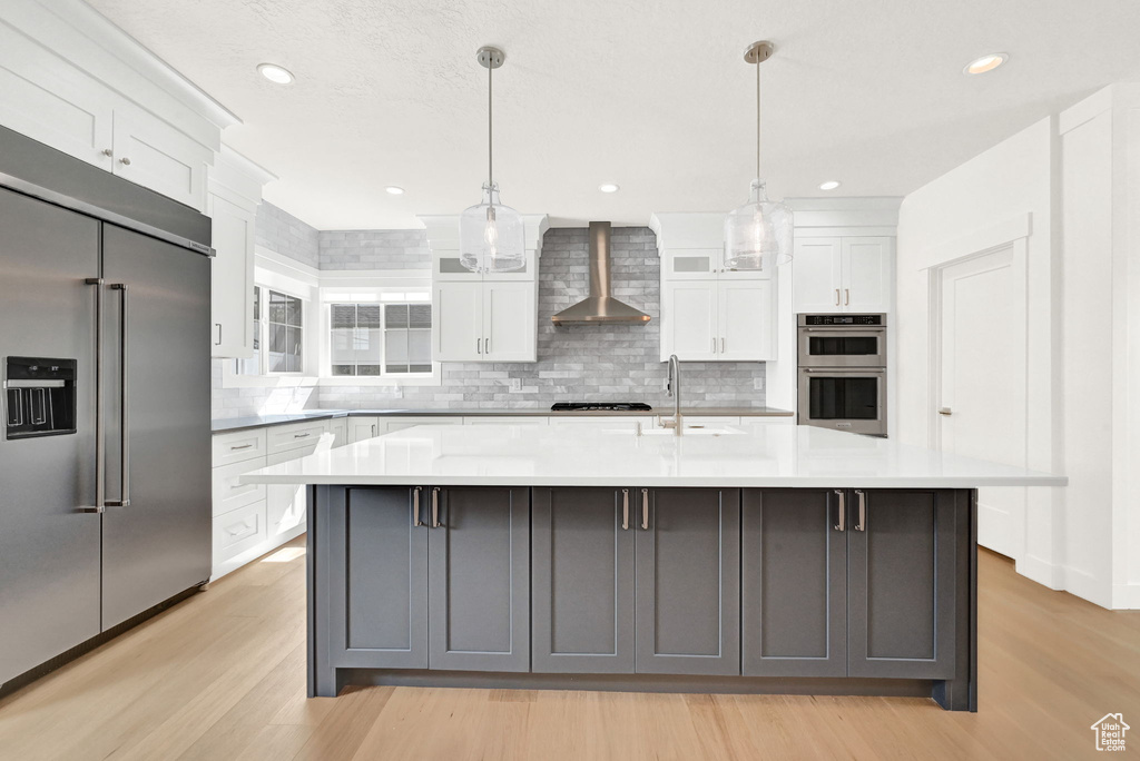 Kitchen with light hardwood / wood-style floors, white cabinetry, wall chimney range hood, stainless steel appliances, and a spacious island