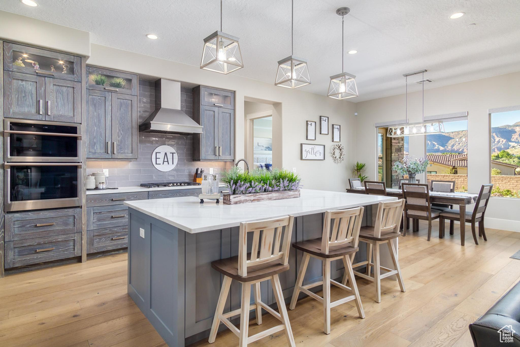 Kitchen featuring light hardwood / wood-style flooring, a kitchen island with sink, wall chimney range hood, and decorative light fixtures