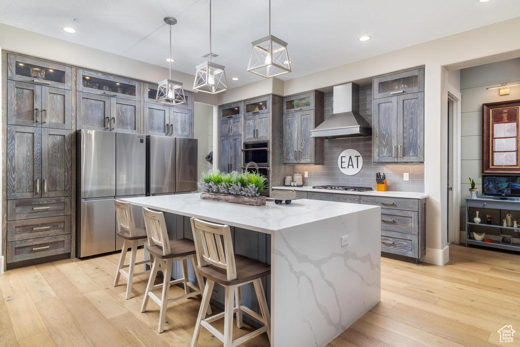 Kitchen with light stone counters, a kitchen island with sink, wall chimney exhaust hood, light hardwood / wood-style flooring, and appliances with stainless steel finishes