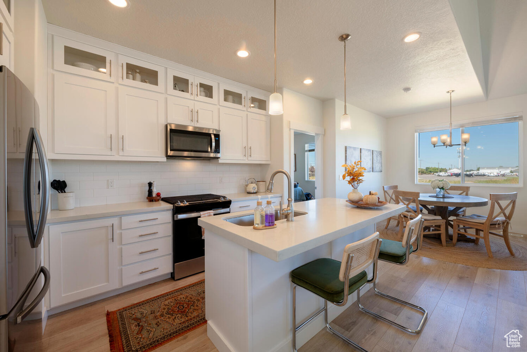 Kitchen featuring light hardwood / wood-style floors, white cabinetry, an island with sink, stainless steel appliances, and a chandelier