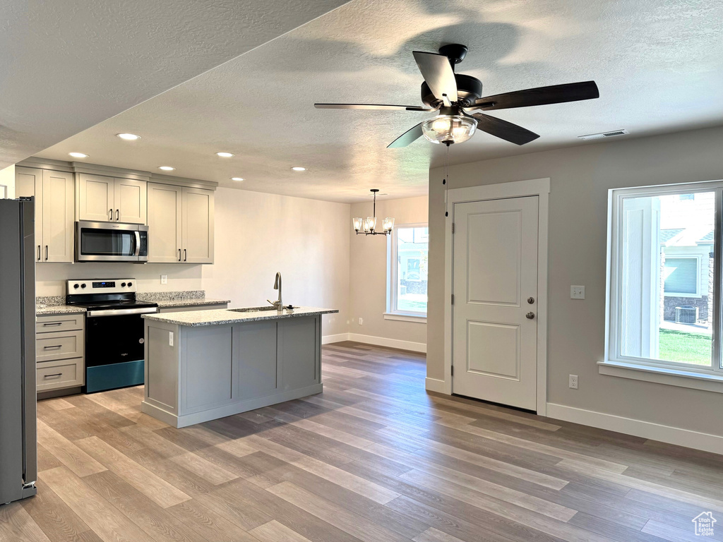 Kitchen with an island with sink, stainless steel appliances, light hardwood / wood-style floors, and ceiling fan with notable chandelier