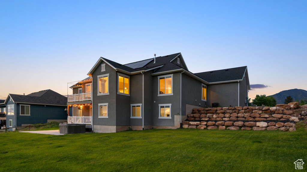 Back of house at dusk with a balcony, a hot tub, a lawn, and stucco siding