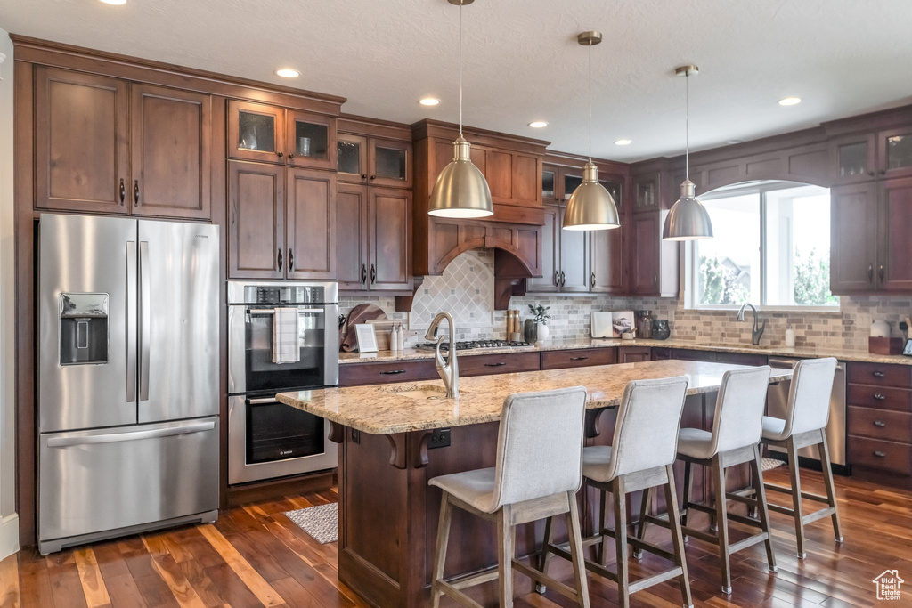 Kitchen featuring light stone counters, a center island with sink, appliances with stainless steel finishes, and dark hardwood / wood-style flooring