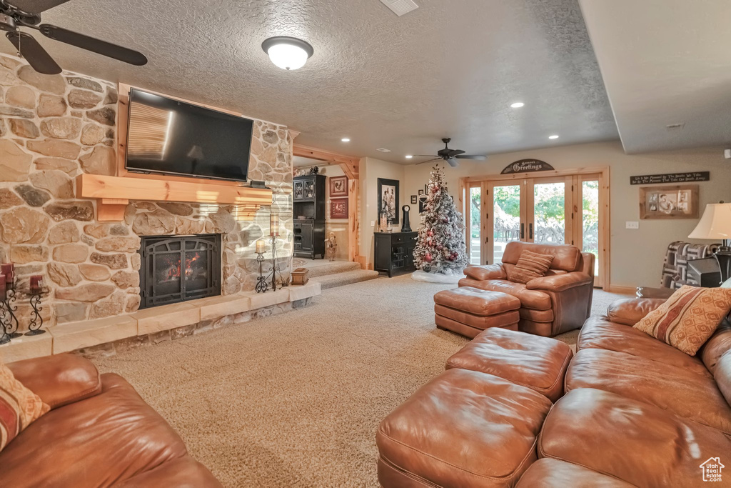 Carpeted living room with a textured ceiling, a stone fireplace, ceiling fan, and french doors