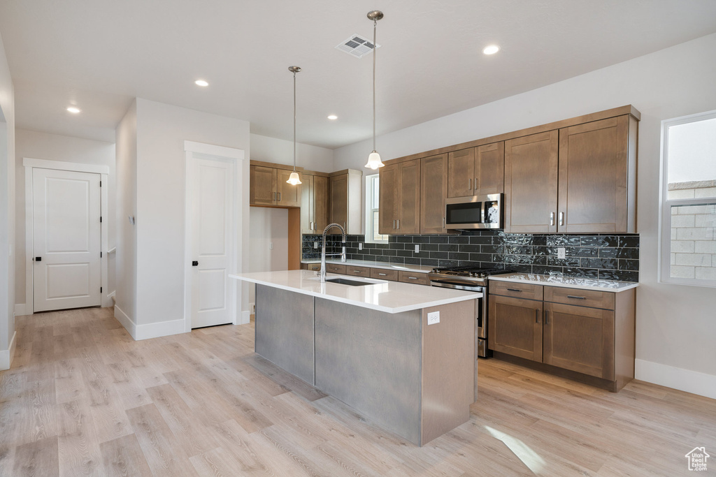 Kitchen with appliances with stainless steel finishes, hanging light fixtures, an island with sink, light hardwood / wood-style flooring, and sink