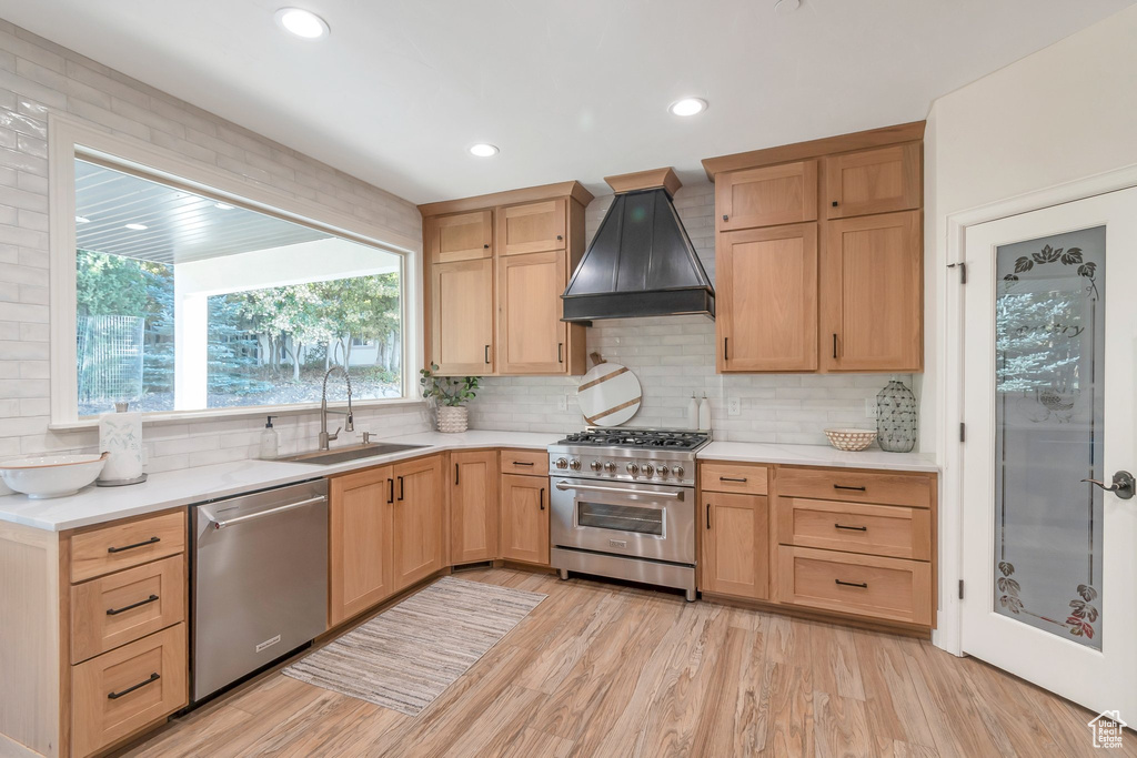 Kitchen featuring custom exhaust hood, sink, appliances with stainless steel finishes, light wood-type flooring, and decorative backsplash