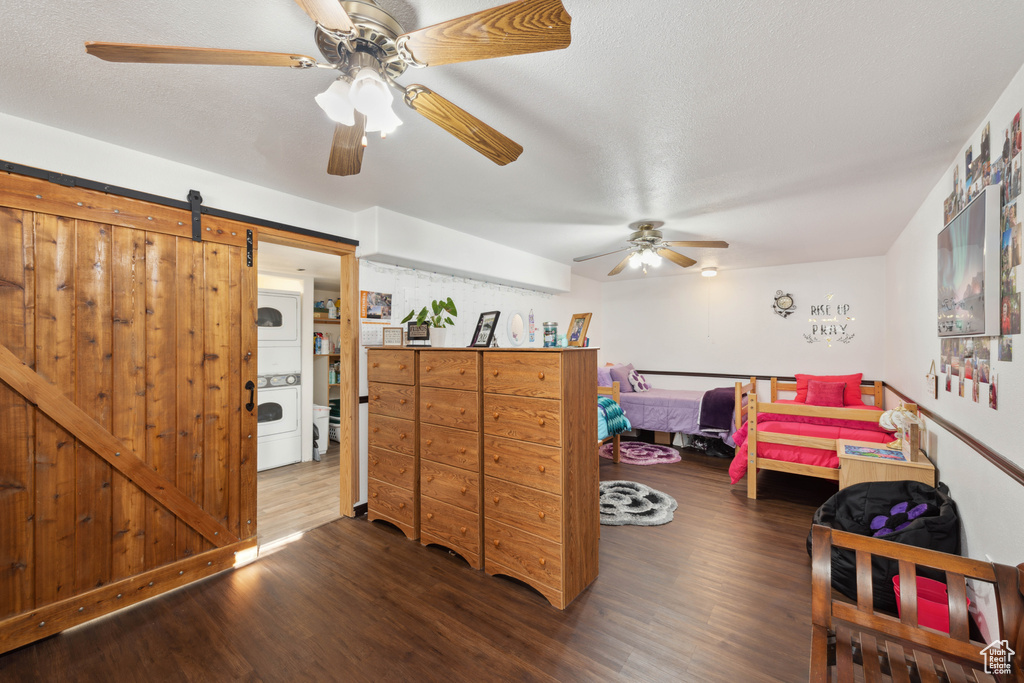 Bedroom featuring a barn door, stacked washing maching and dryer, dark hardwood / wood-style flooring, and ceiling fan