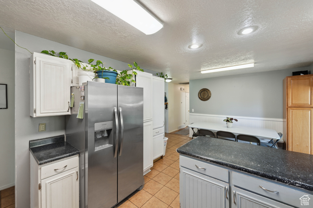 Kitchen with a textured ceiling, stainless steel fridge with ice dispenser, white cabinetry, light tile patterned floors, and dark stone countertops